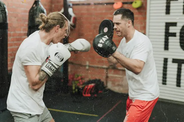 Dos hombres entrenando boxeo con guantes y manoplas en un gimnasio de paredes de ladrillo