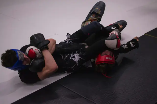 Dos luchadores con casco y guantes forcejeando en el suelo sobre el tatami